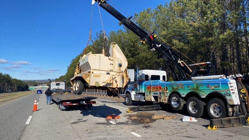 Tow truck lifting overturned beige truck on highway shoulder beside scattered debris and cones