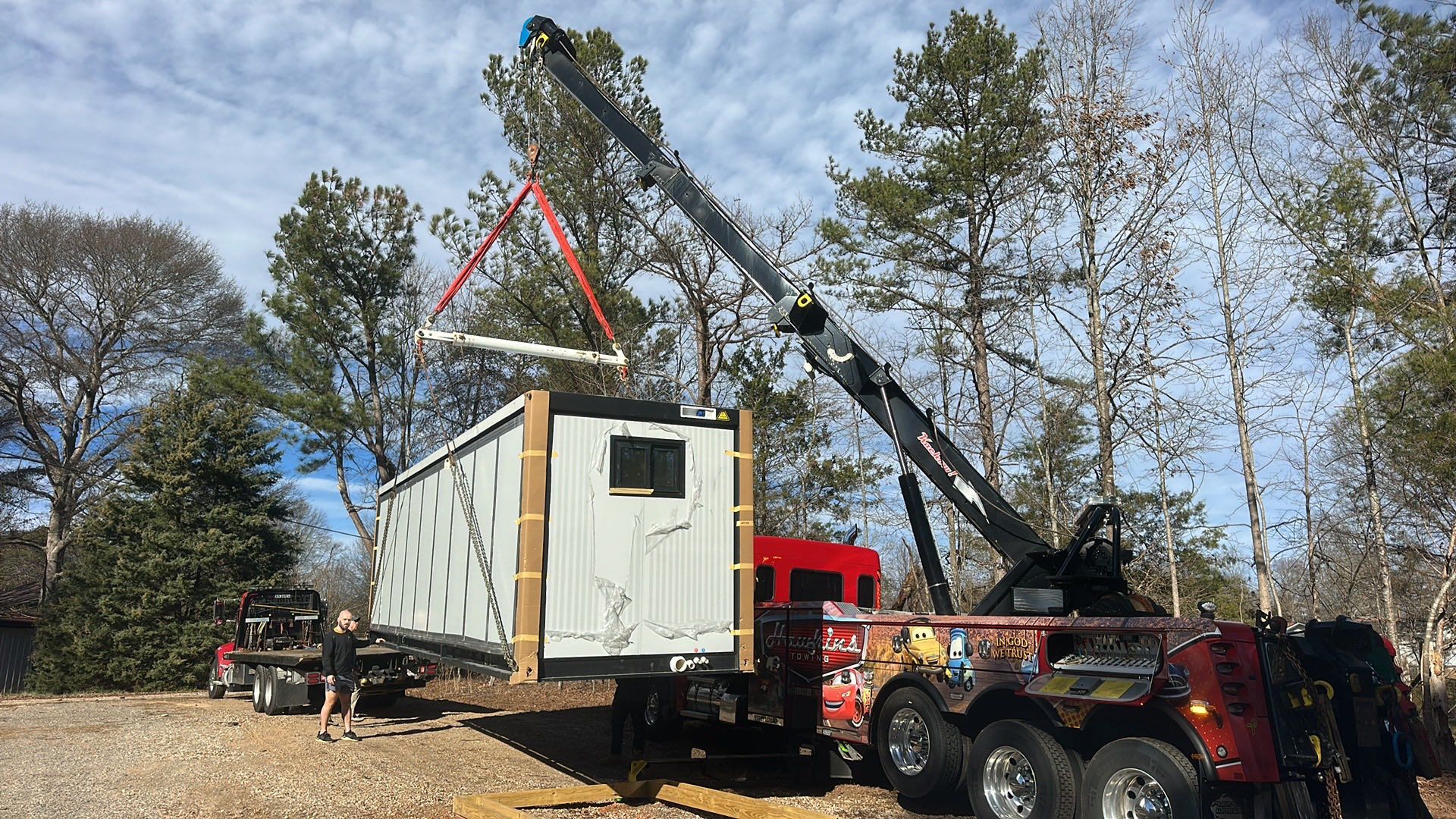 Crane lifts a white shipping container onto a truck in a wooded outdoor area.