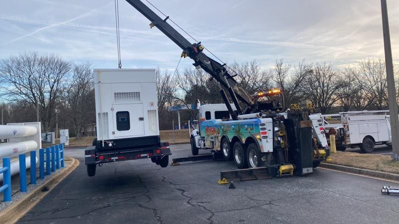 Tow truck lifting a white box truck on a street beside blue railings, with a crane arm extended.
