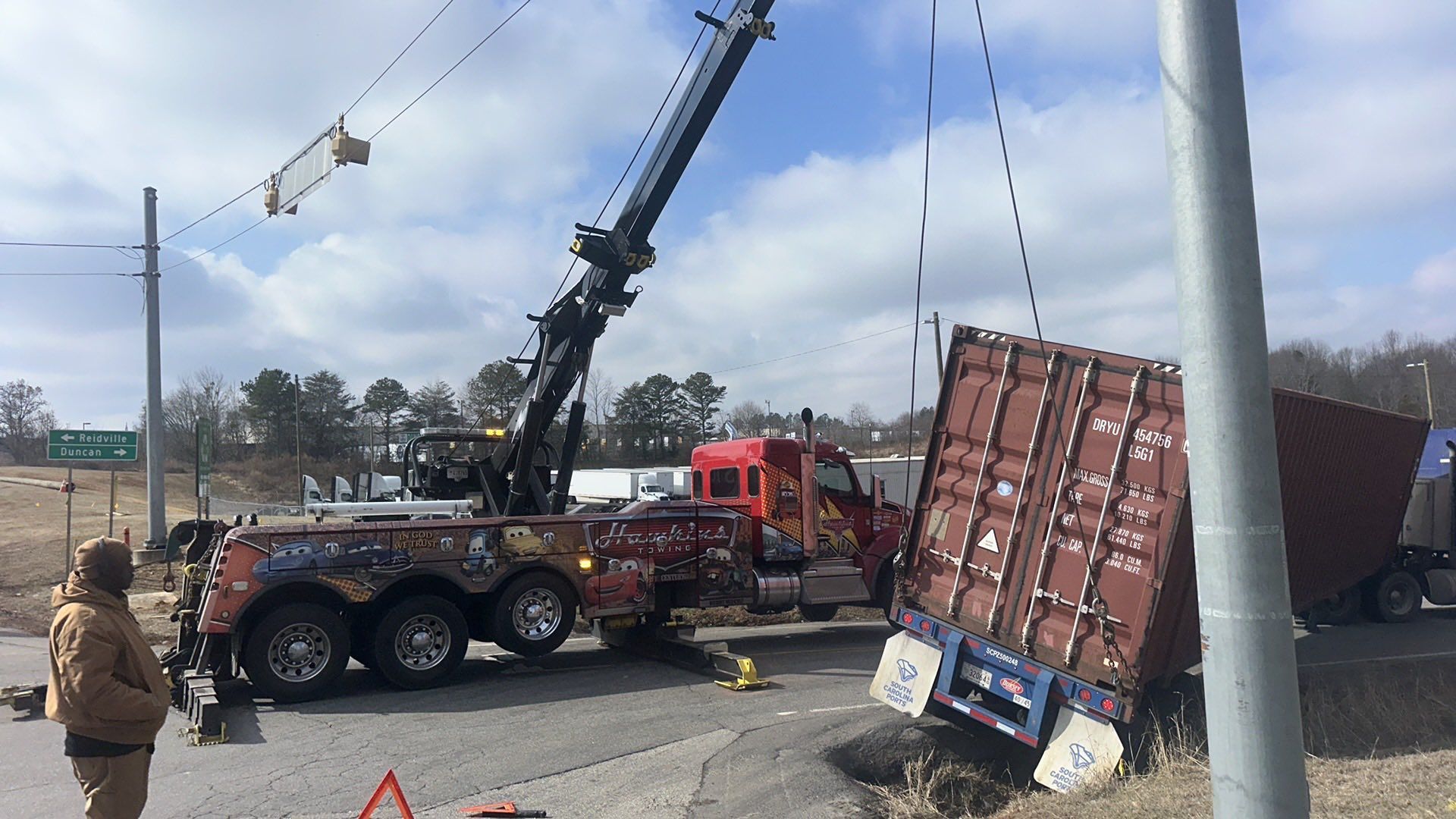 Tow truck lifting overturned red trailer beside a damaged semi on a roadside under a blue sky