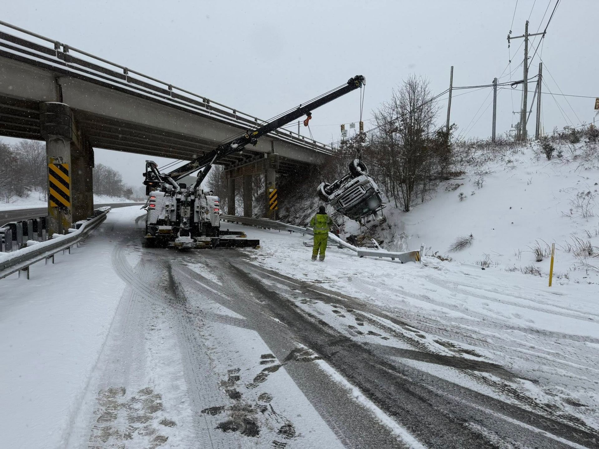 Tow truck recovering a car wreck from a snowy roadside under an overpass, with a worker nearby.