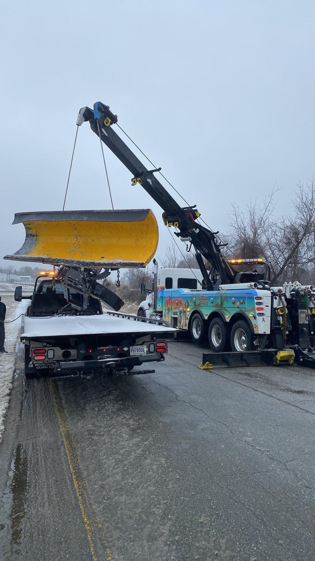 Tow truck lifting a yellow snow plow blade from a pickup on a wet road in light rain