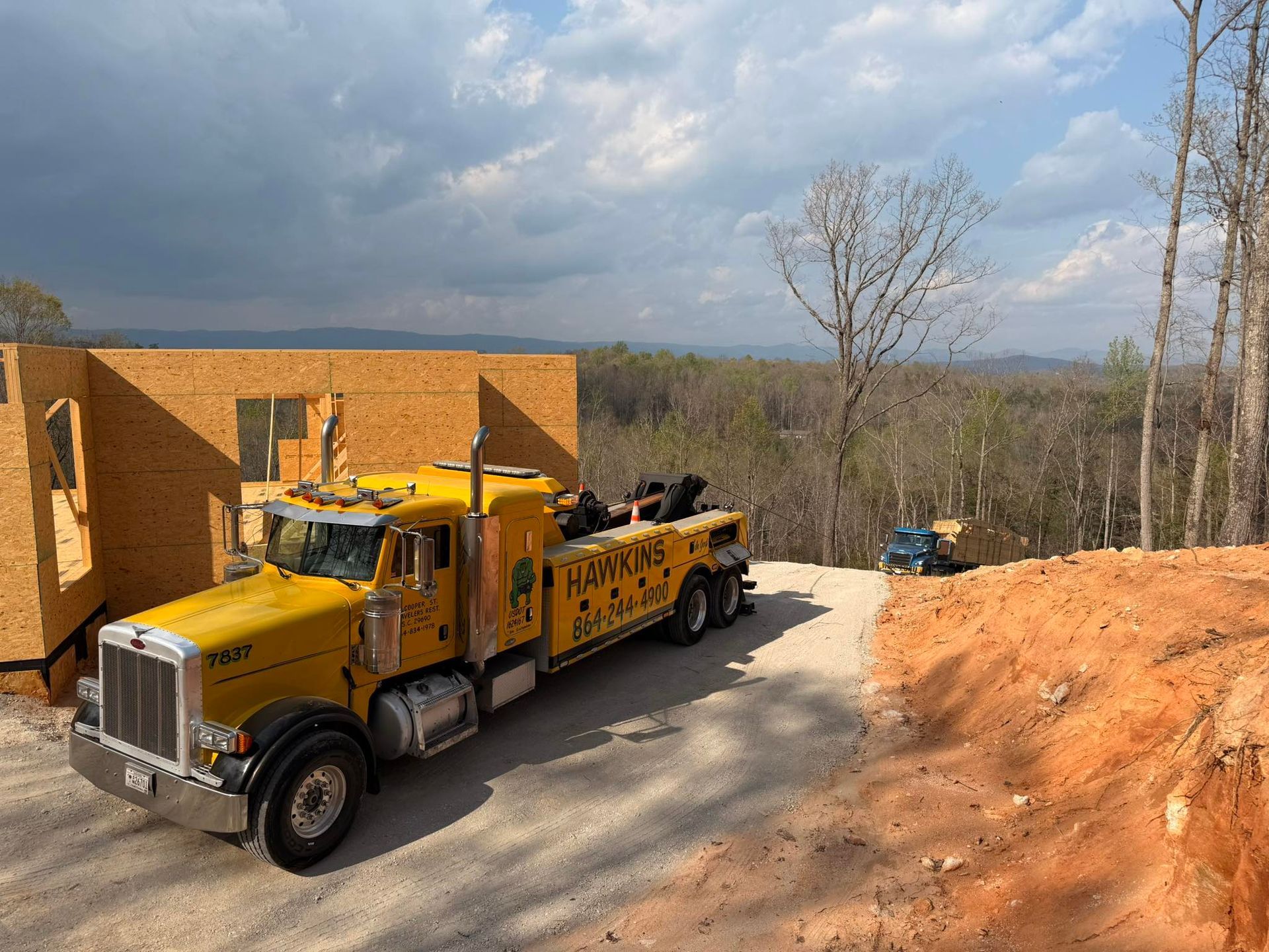 Yellow tow truck beside a brick building on a dirt construction site under a cloudy sky