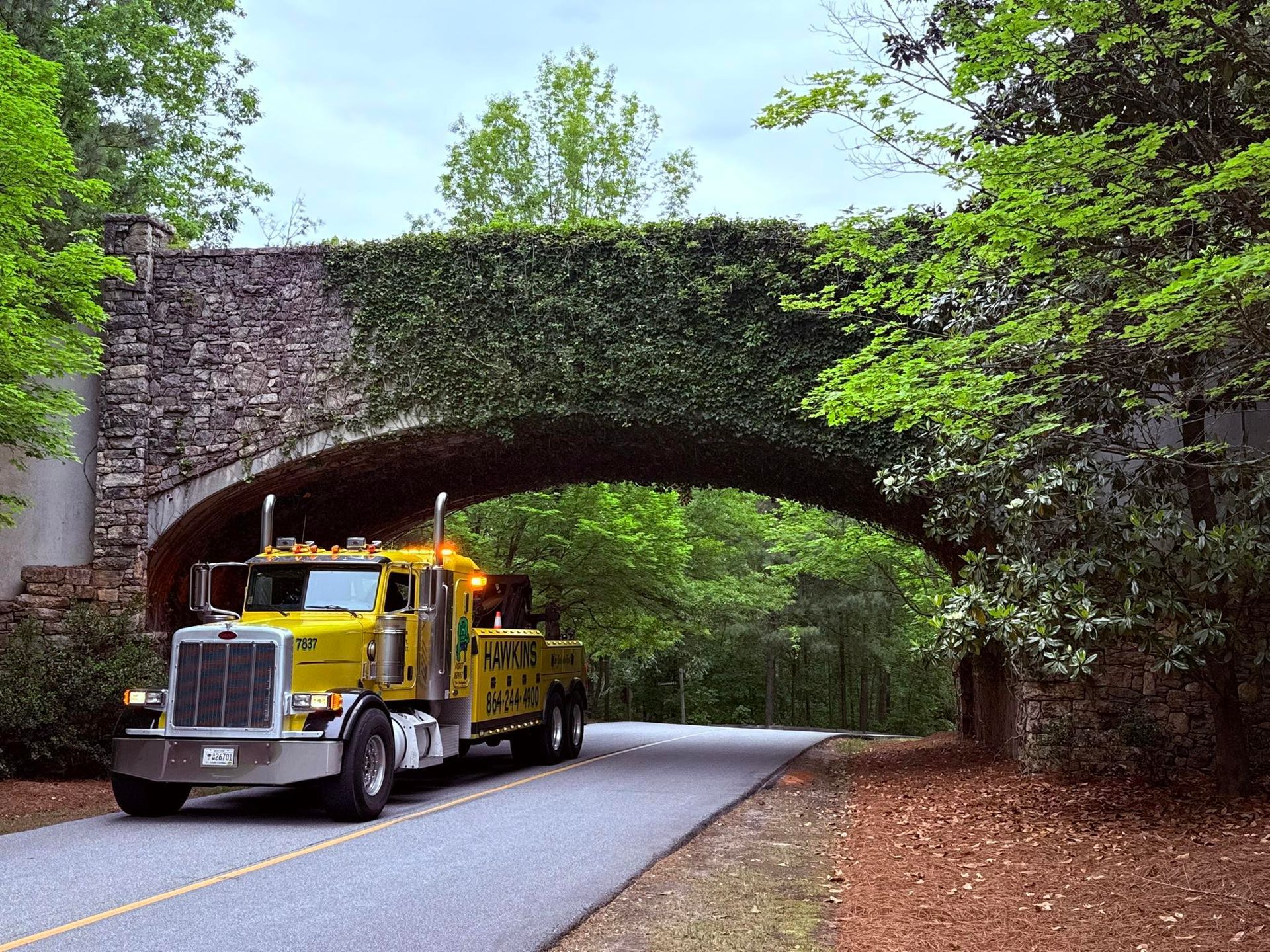 Yellow tow truck drives under a stone arch on a tree-lined road.