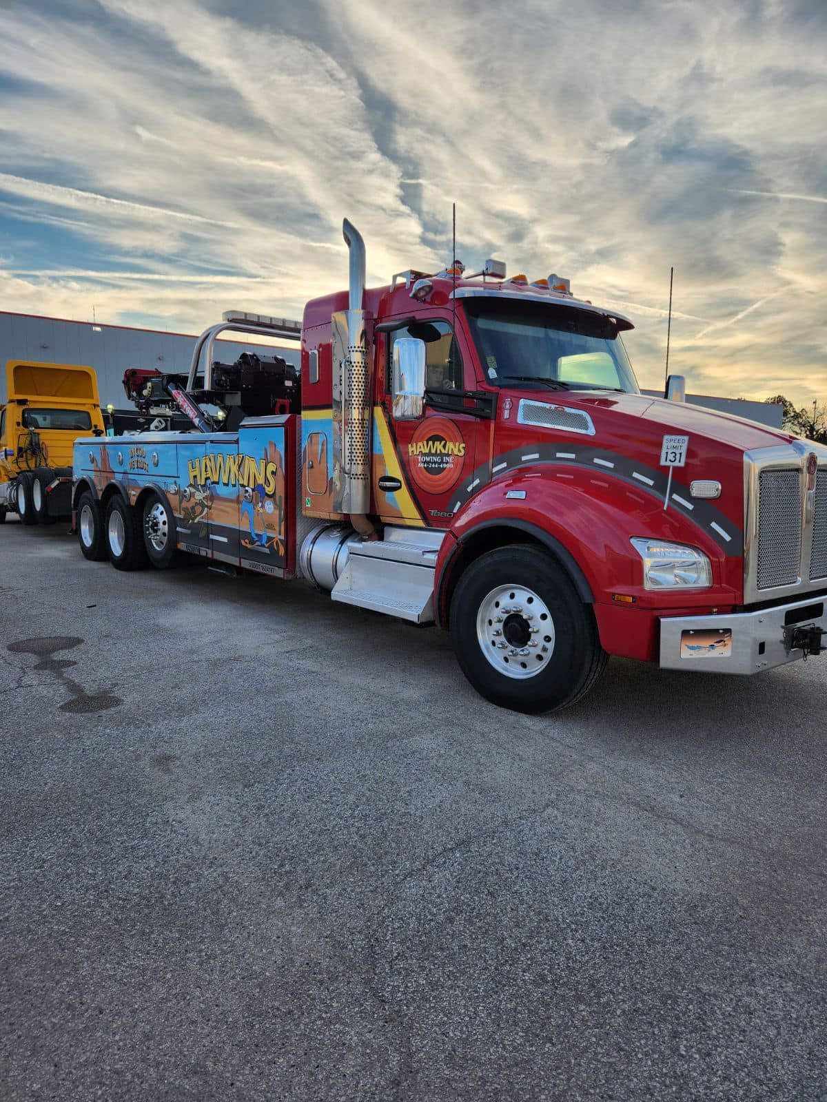 Red tow truck with colorful side graphics parked on gravel at sunset, with another truck behind it.
