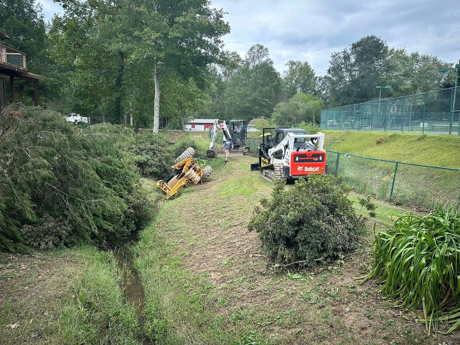 Small excavator and mini skid steer clearing a grassy backyard path with cut branches and shrubs.