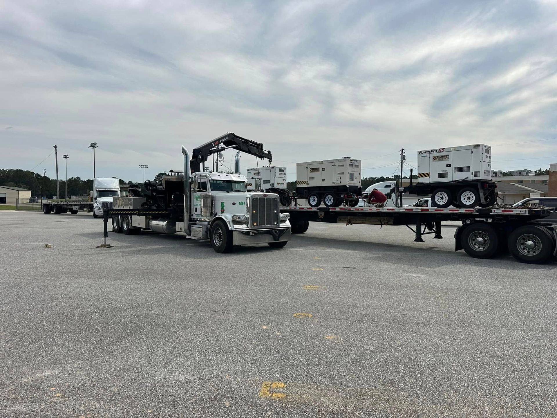 White semi-truck with crane loading flatbed trailers in a gravel lot under cloudy sky
