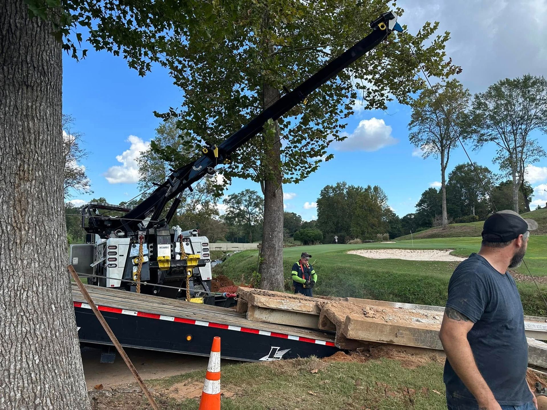 Tow truck on a dirt road lifting a car by a park, with workers and orange cones nearby