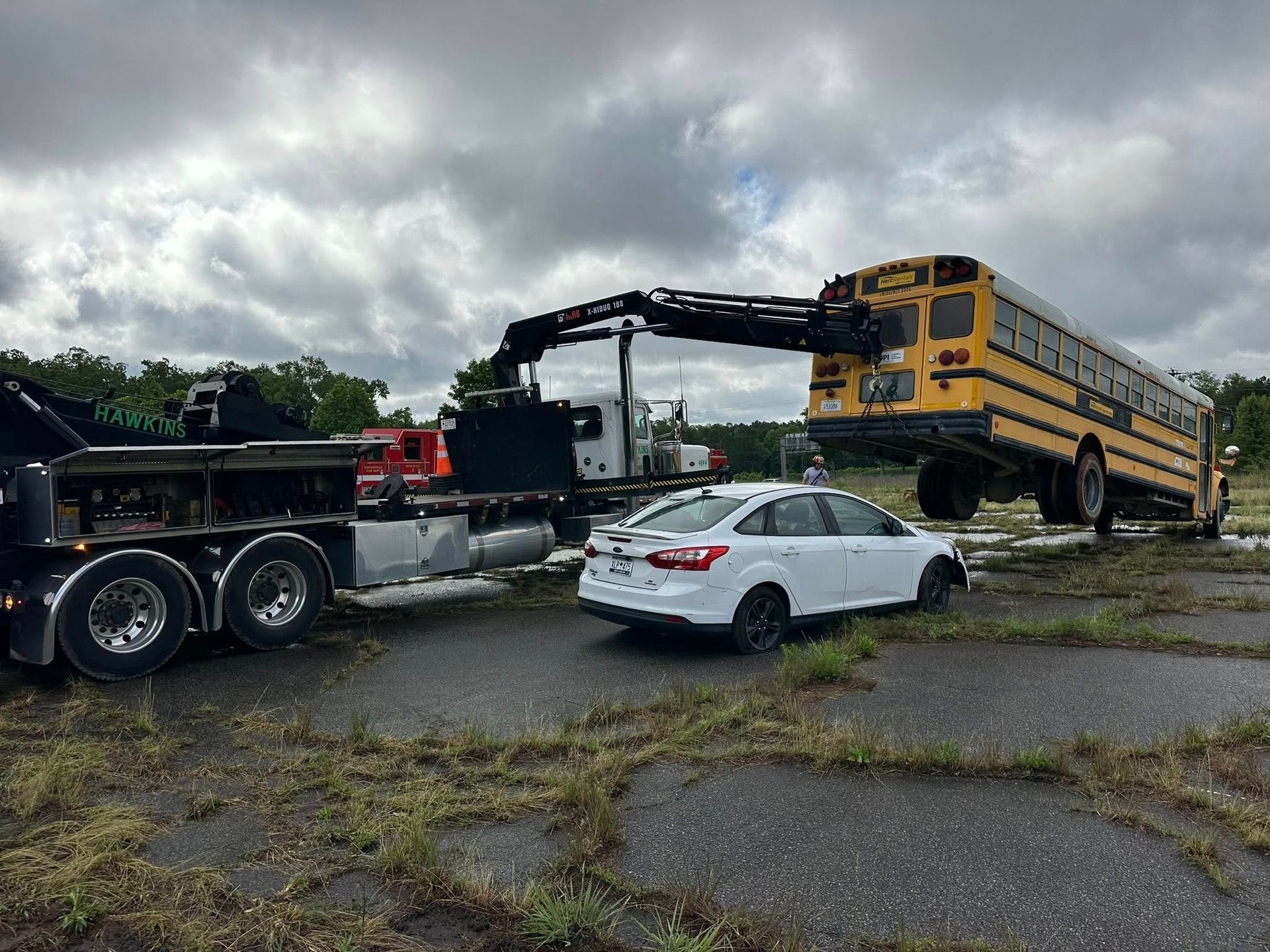 Tow truck lifting a yellow school bus beside a white sedan in a cloudy parking lot