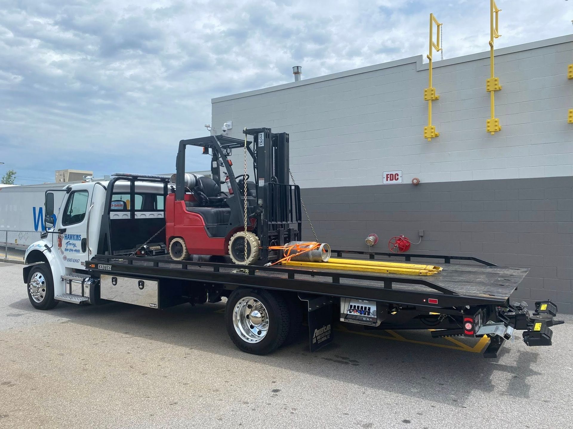Tow truck with forklift loaded on flatbed parked beside a building