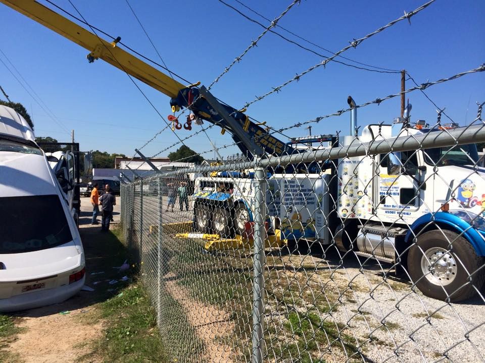 Tow truck lifting a white truck beside a chain-link fence under a clear blue sky