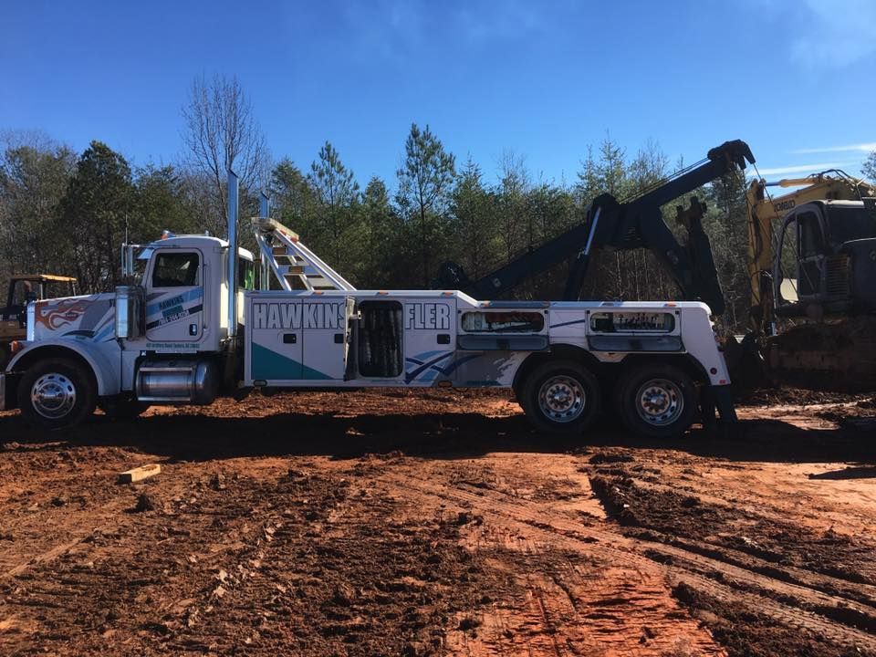 White tow truck with boom on red dirt road beside a ditch, with another truck and trees in the background