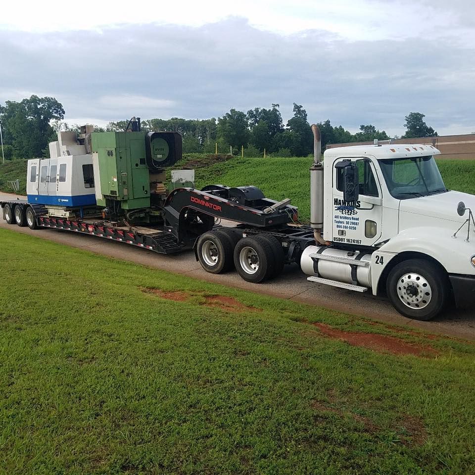 White tow truck hauling a large green-and-white machine on a flatbed along a rural road