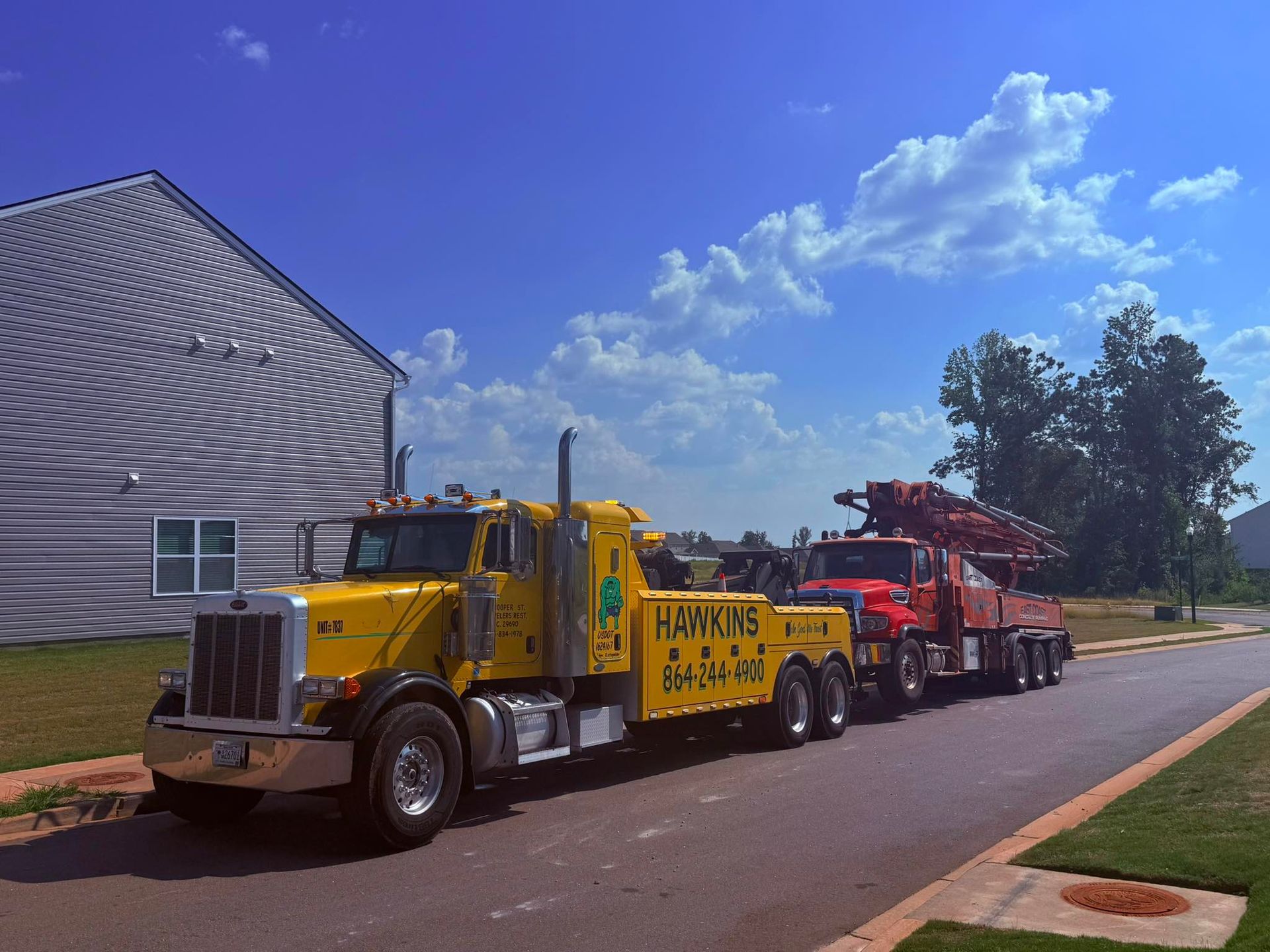 Yellow tow truck hauling red heavy equipment on a road beside a white building under a blue sky.