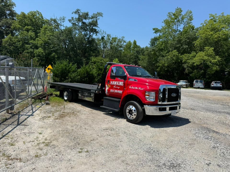 Red flatbed tow truck parked on a gravel lot beside a chain-link fence and trees.