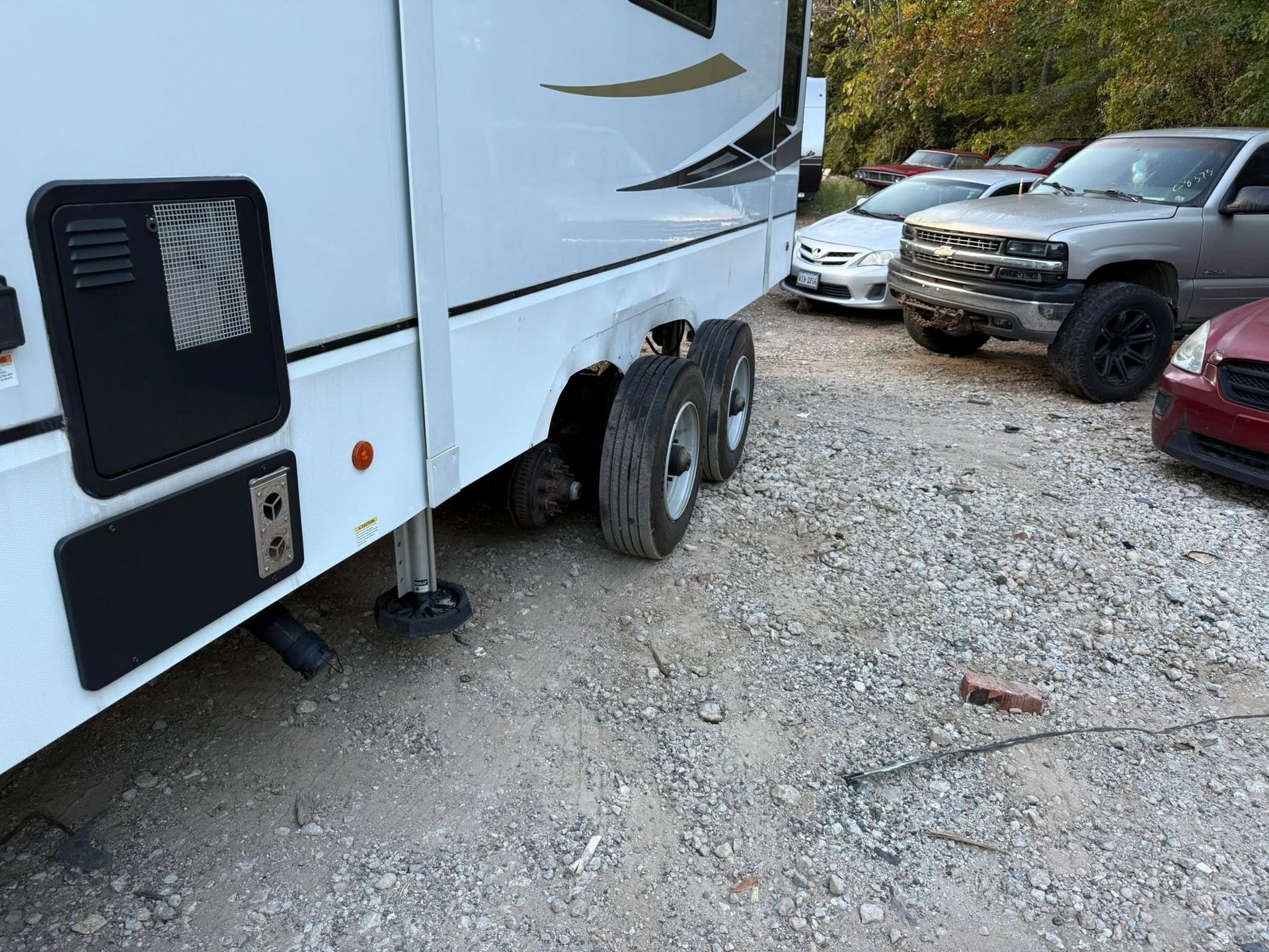 White RV trailer parked on gravel beside several trucks and cars in a lot.