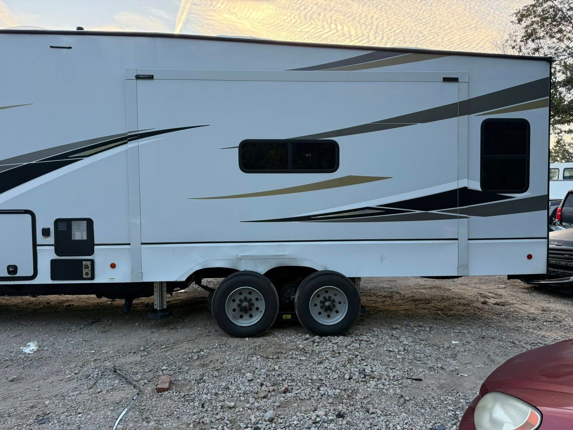 White dual-axle travel trailer parked on gravel, with black and gray swoosh graphics.