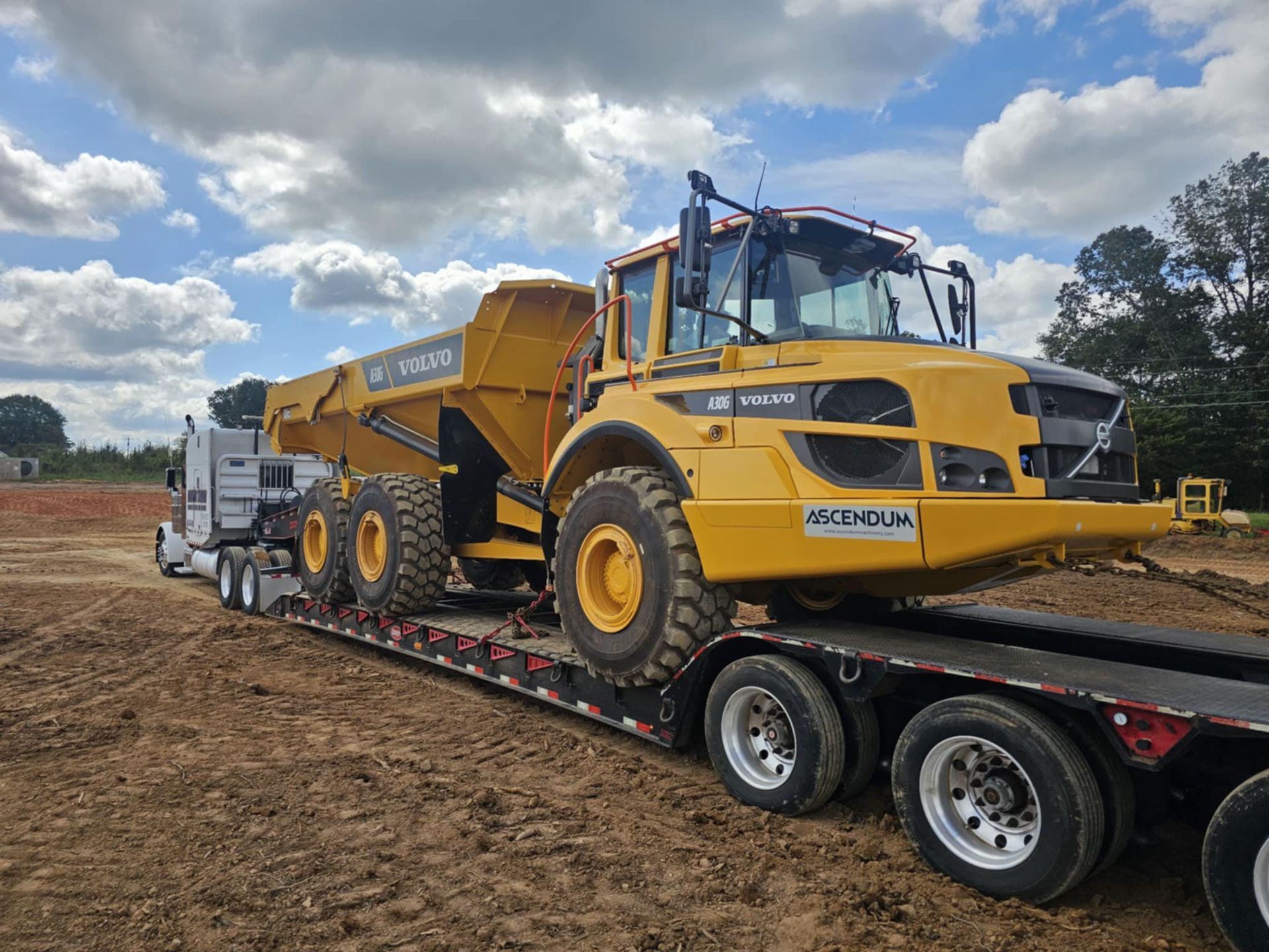 Yellow construction dump truck on a flatbed trailer in a dirt field under a cloudy sky