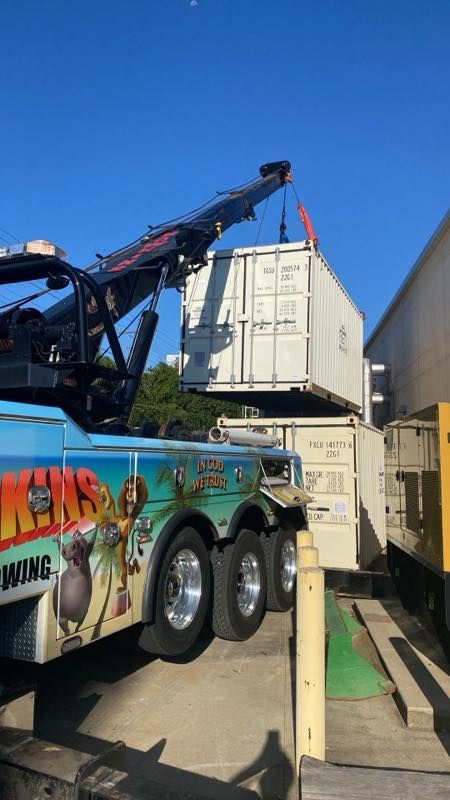 Tow truck lifting a white shipping container beside a building under a clear blue sky