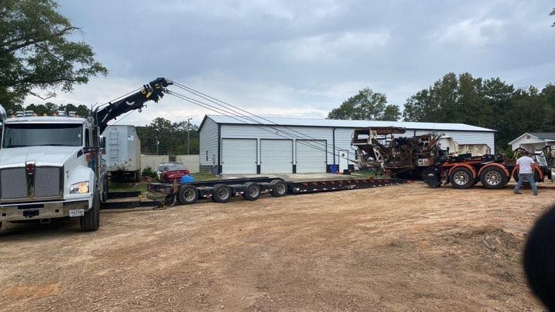 Tow truck hauling a large red tractor beside a white garage on a gravel lot under cloudy skies