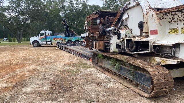 Tracked demolition machine hauling a white RV on a flatbed trailer in a muddy lot