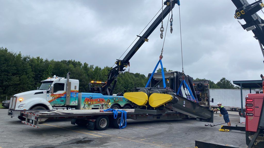 Tow truck lifting a black car from a flatbed trailer in a gravel lot, with a crane and truck nearby