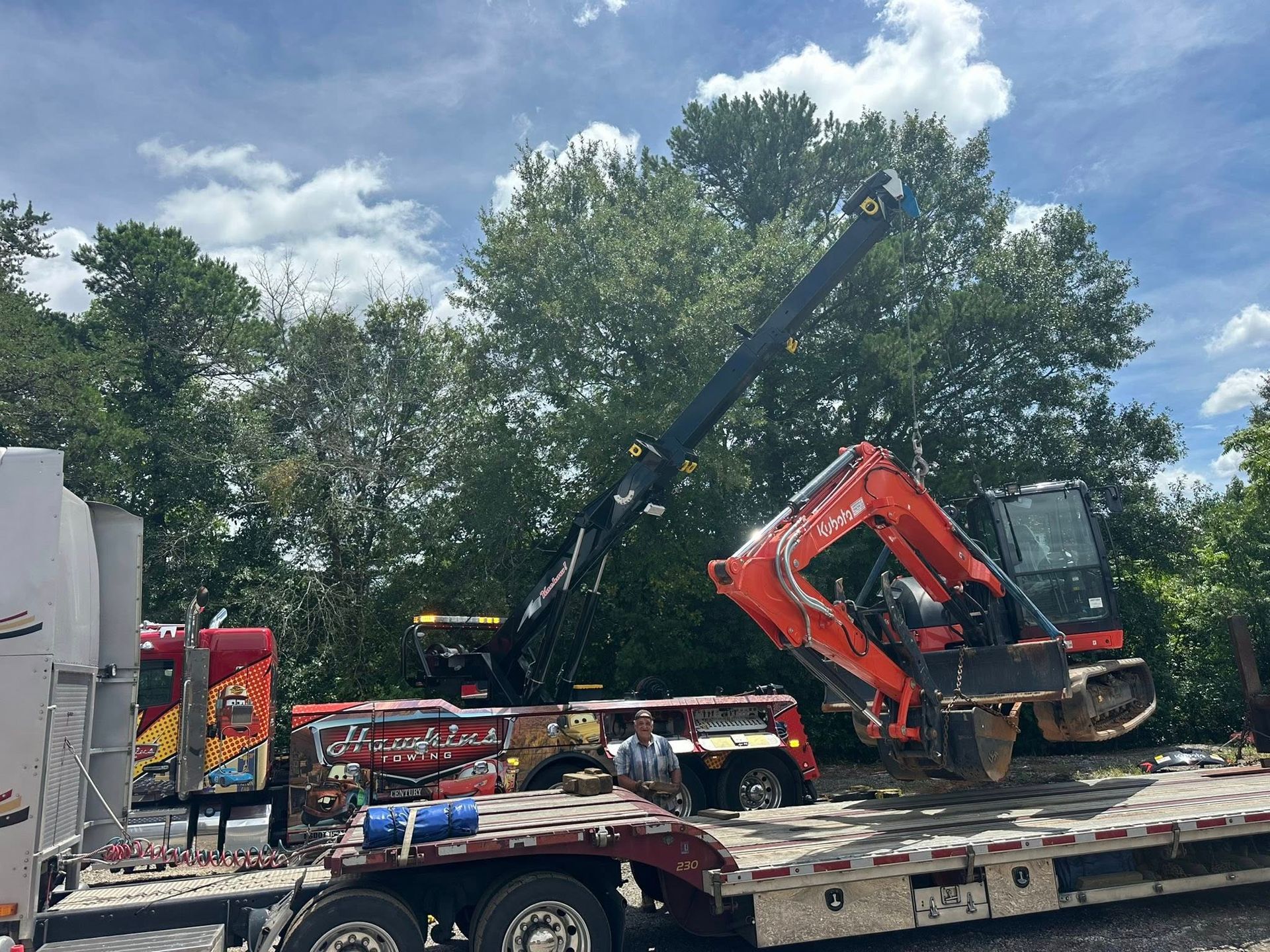 Red excavator being loaded onto a flatbed trailer with a crane in a tree-lined lot