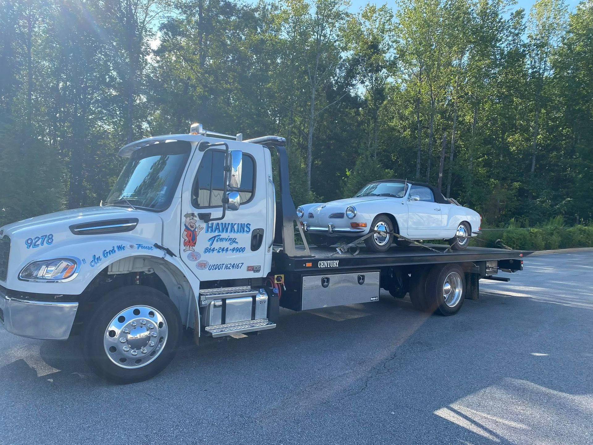 Tow truck carrying a white classic car on a flatbed, parked beside trees in daylight.