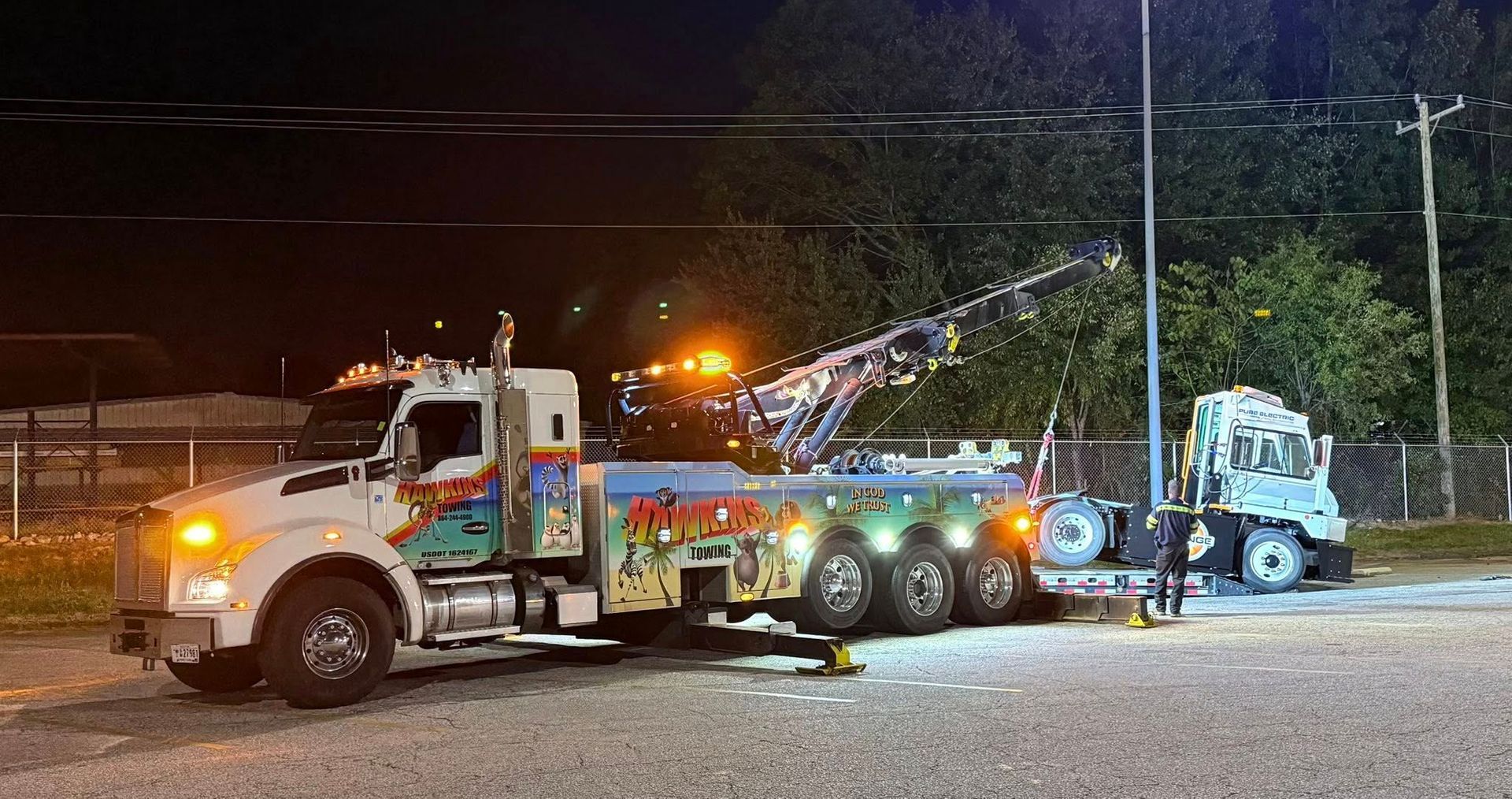 Tow truck towing a blue car at night on a roadside, with flashing lights and a worker nearby