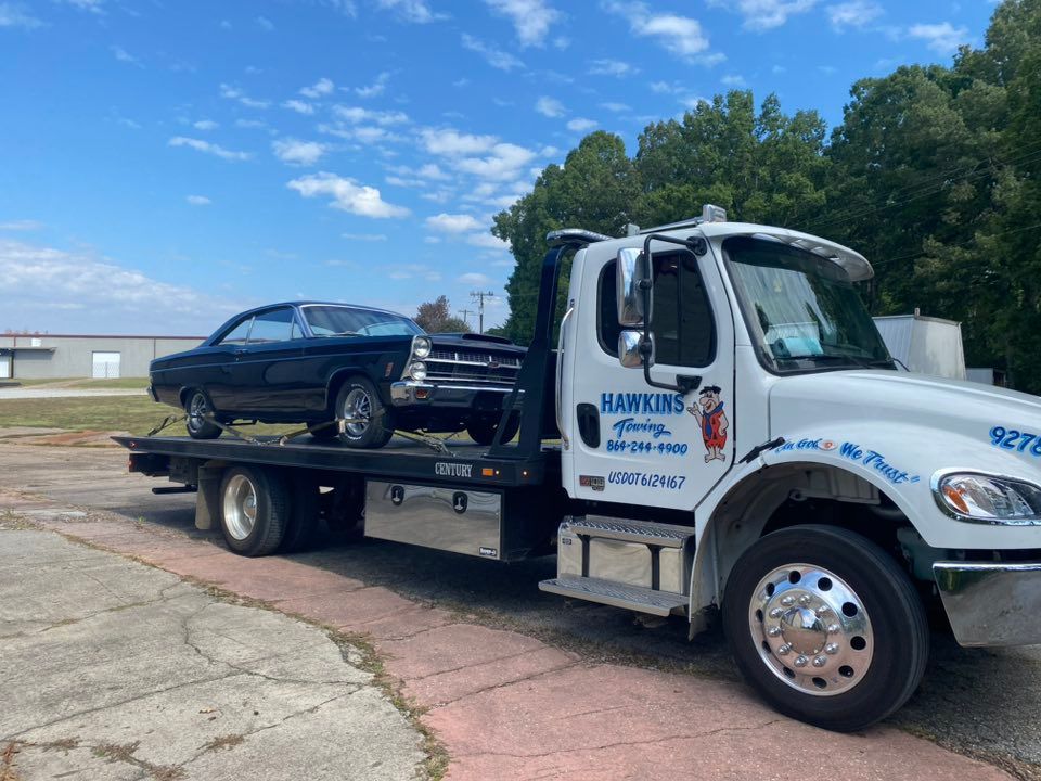 Blue classic car on a flatbed tow truck parked outdoors under a bright sky.