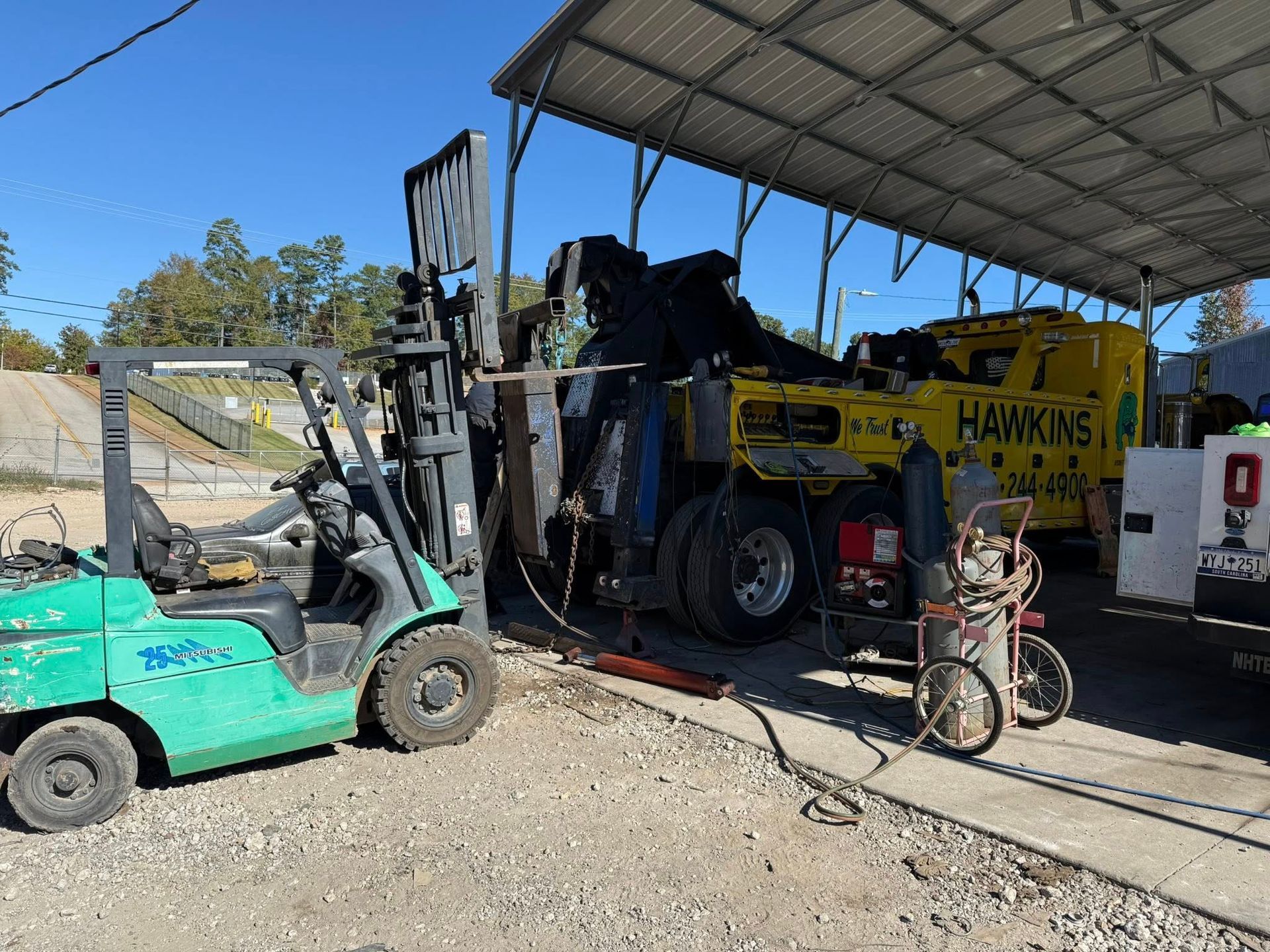 Forklift beside a yellow Hawkins truck under a metal shed, with a green cart and bicycle in front