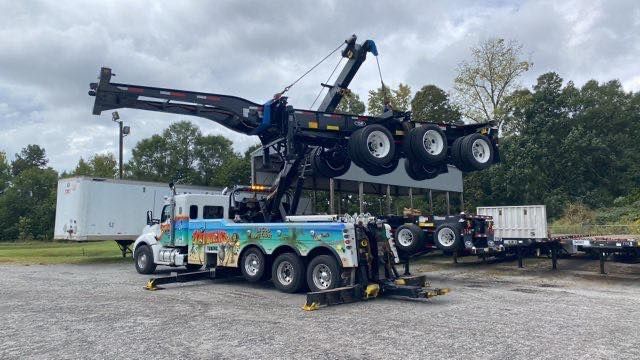 Tow truck lifting a large black semi-truck chassis in a gravel lot under cloudy skies