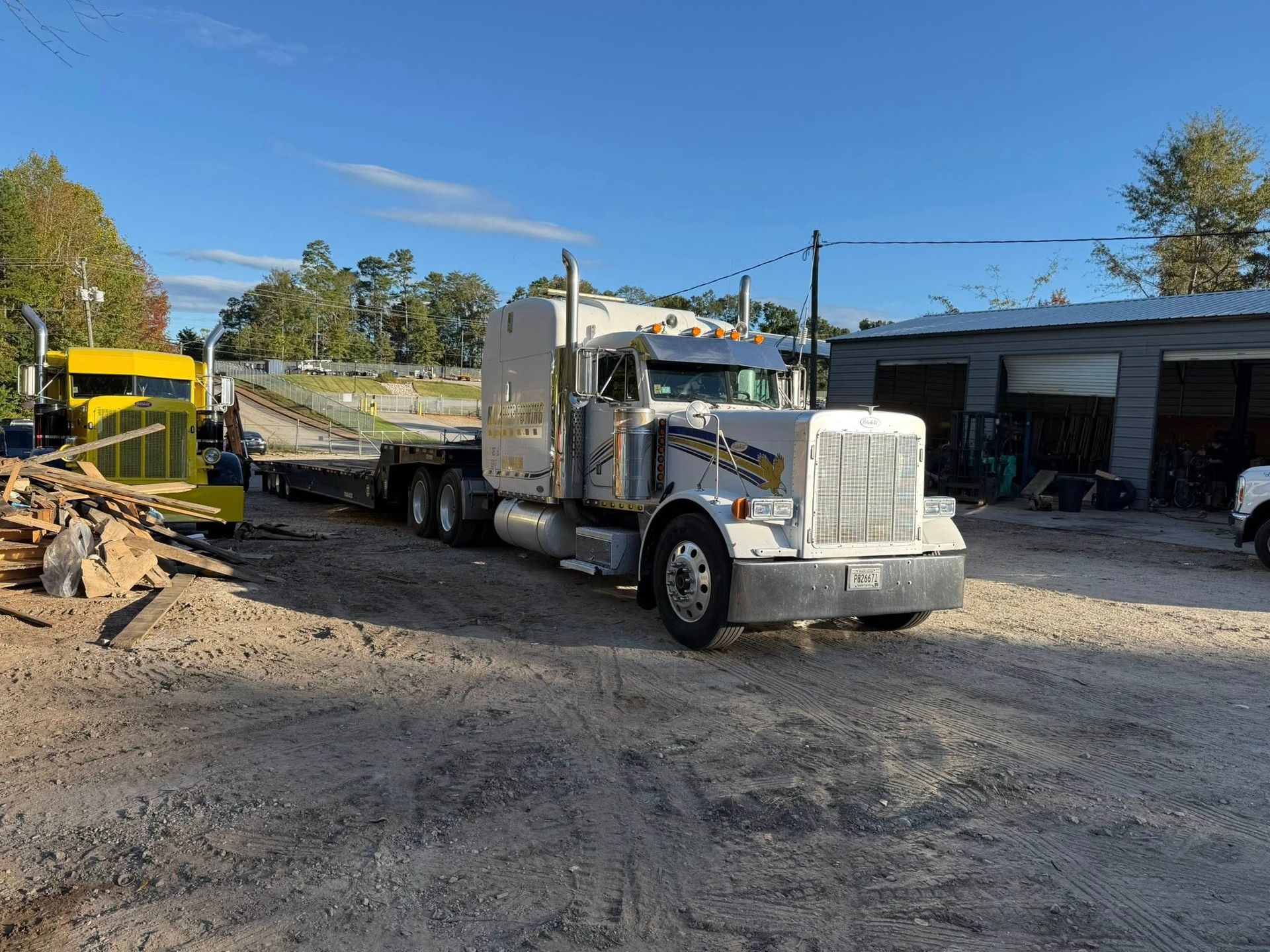 White semi truck with flatbed trailer parked in a gravel yard beside equipment and a shed