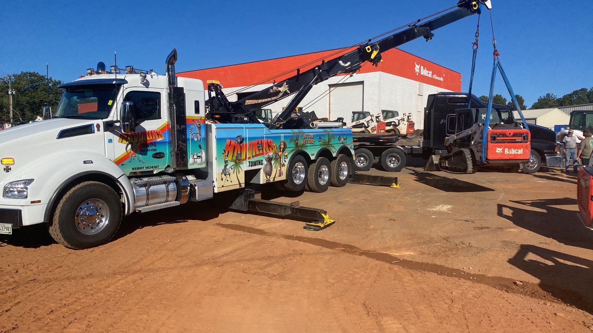 Tow truck with raised crane lifting a white truck in a dirt lot under a clear blue sky