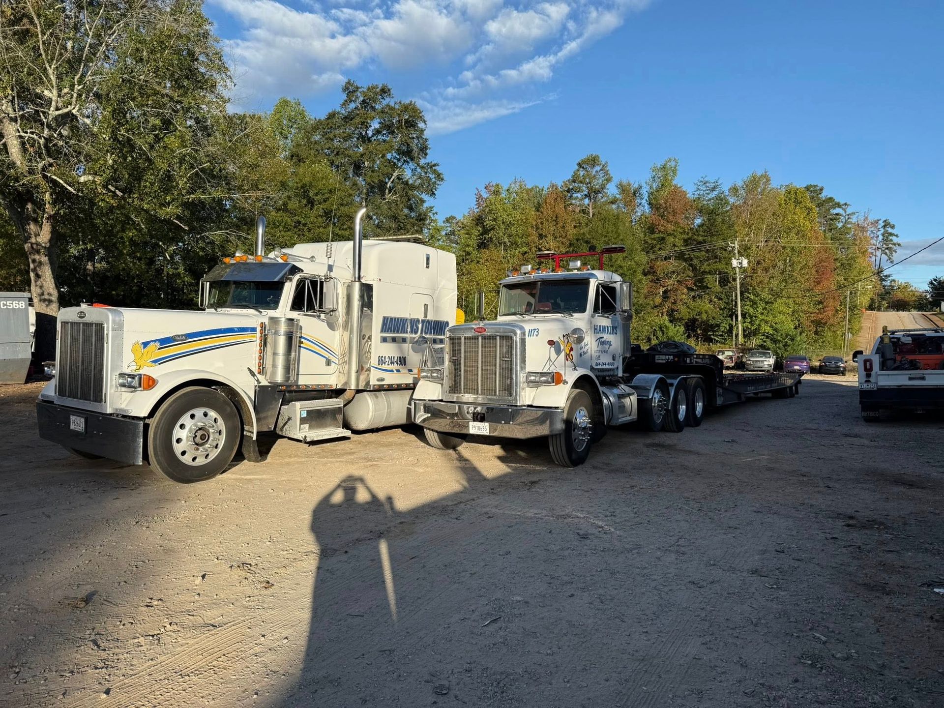Two white semi trucks parked on a gravel lot with trees and blue sky in the background