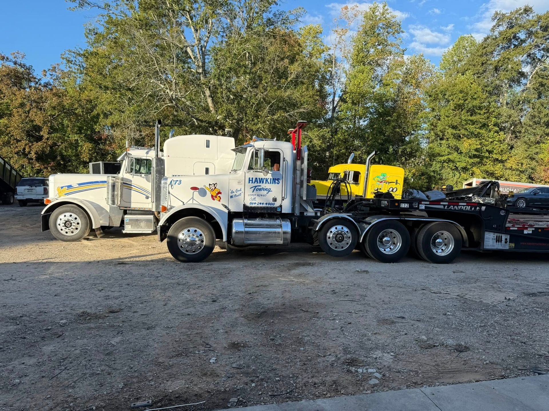 White semi truck with flatbed trailer parked on gravel under trees, with a yellow machine on the trailer.