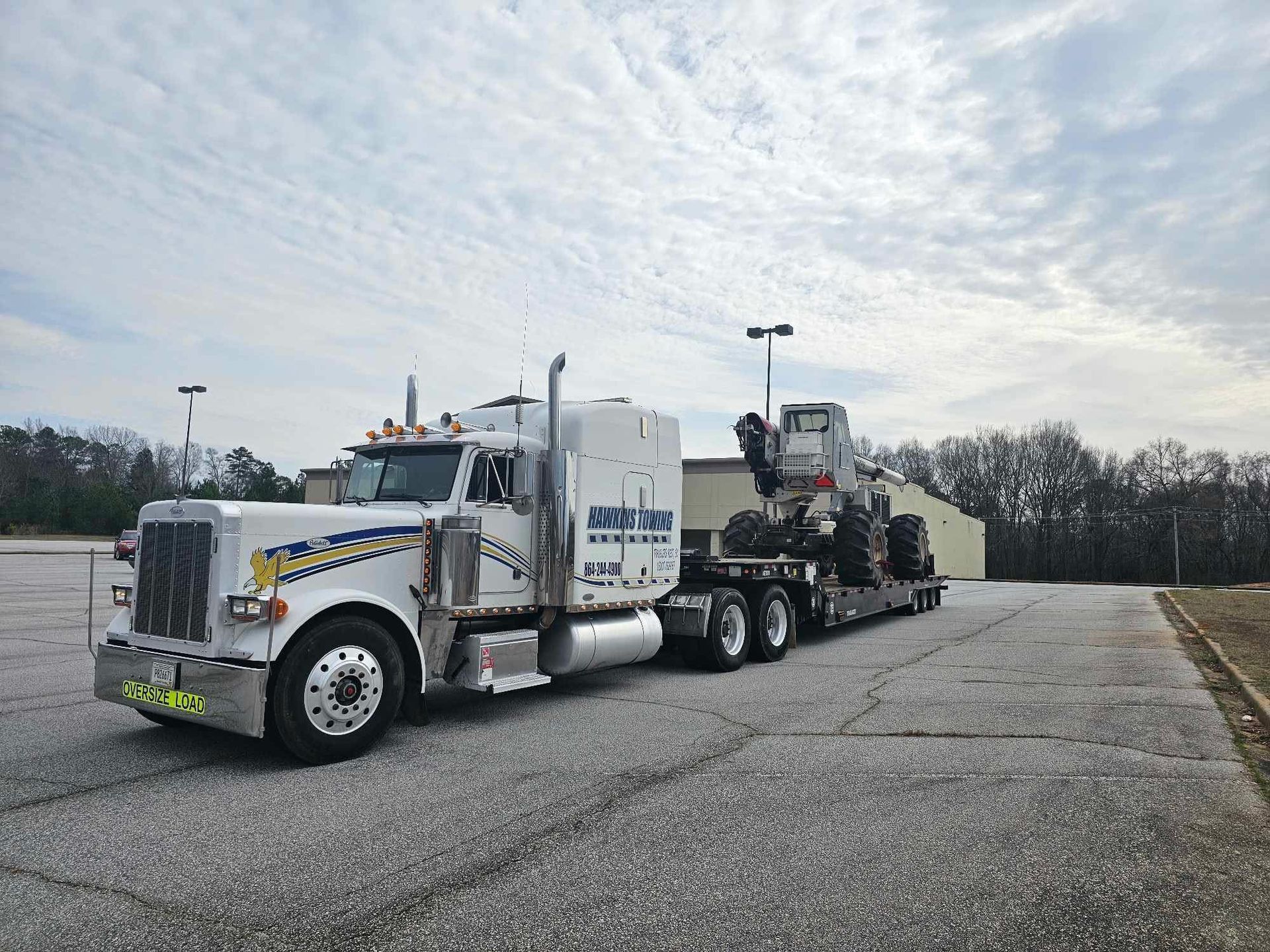 White tow truck hauling a loaded flatbed trailer in a parking lot under a cloudy sky