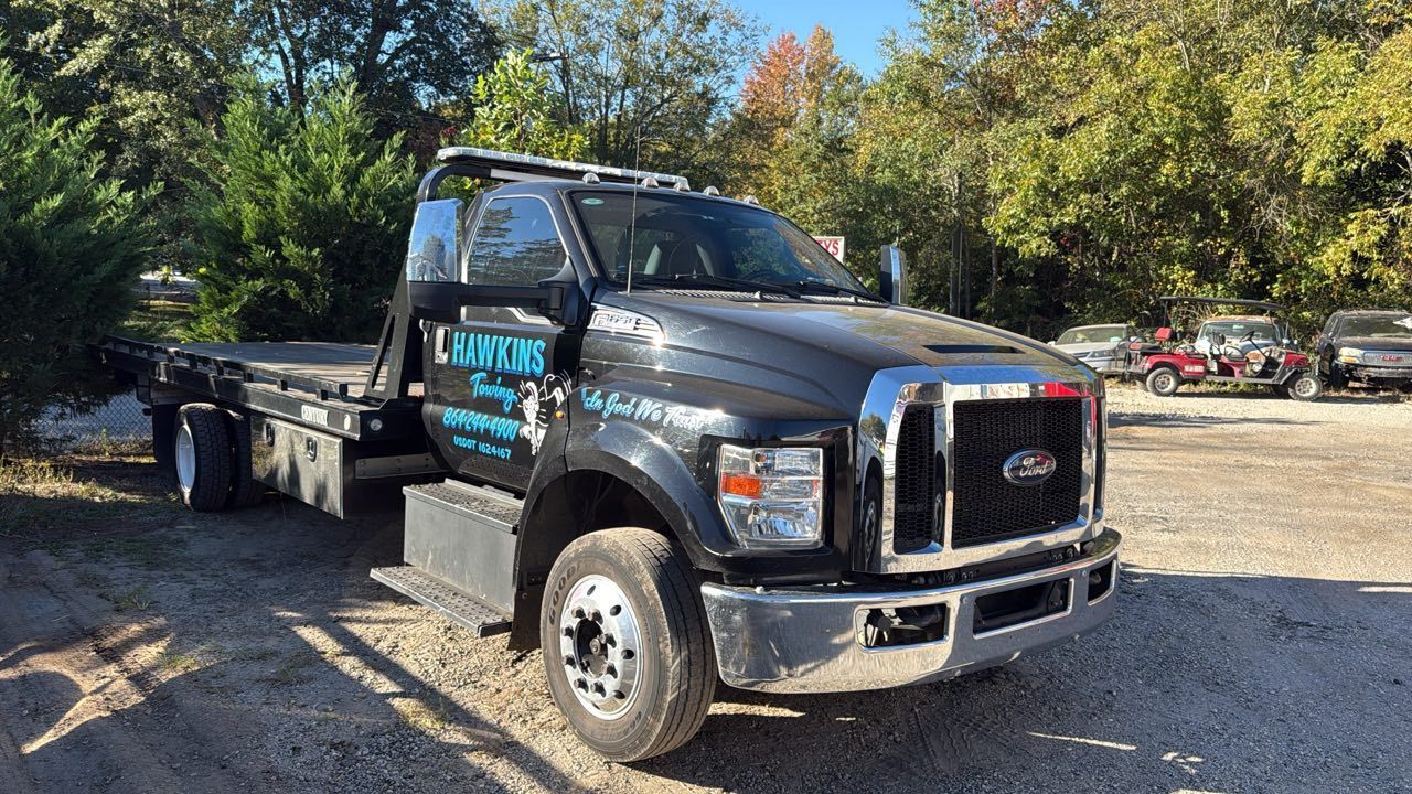 Black flatbed tow truck parked outdoors on a sunny gravel lot with trees in the background
