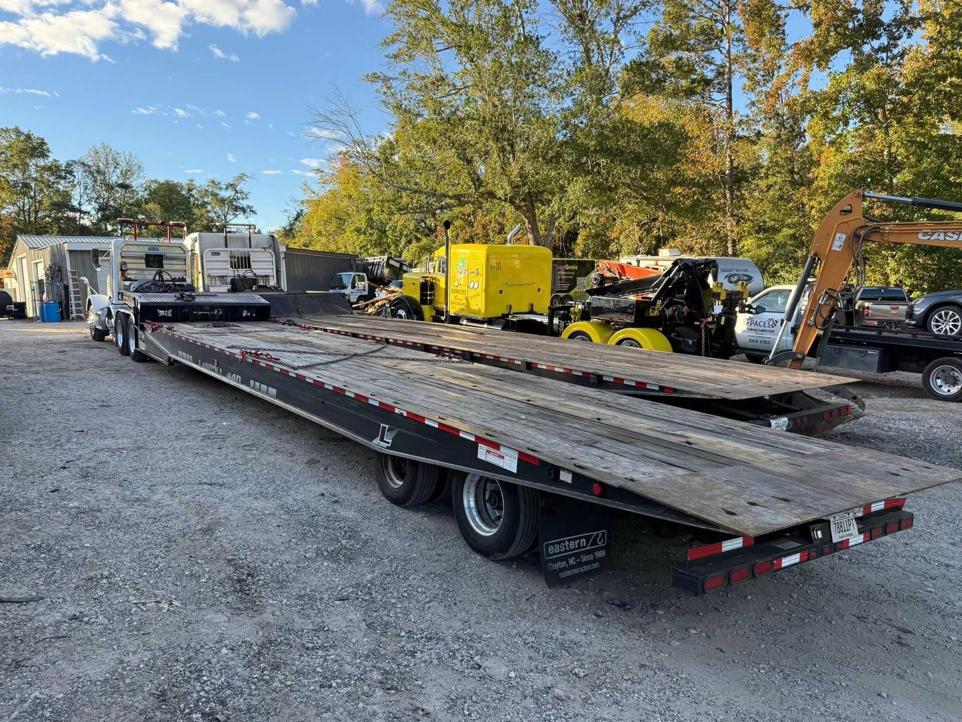 Flatbed trailer loaded with large metal beams in a gravel lot, with trucks and equipment in the background