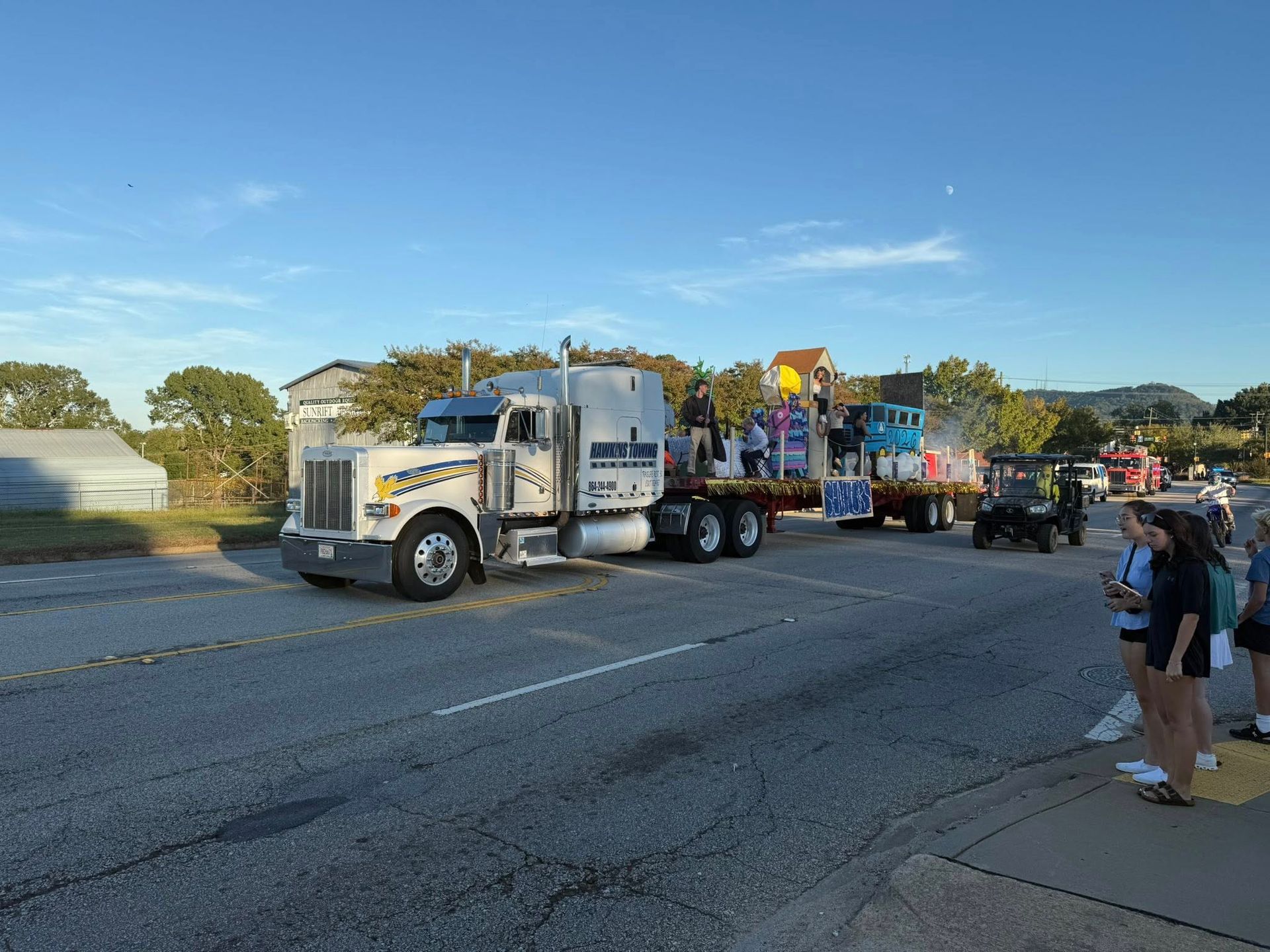 Tow truck hauling a decorated float and vehicles on a roadside, with spectators nearby at sunset