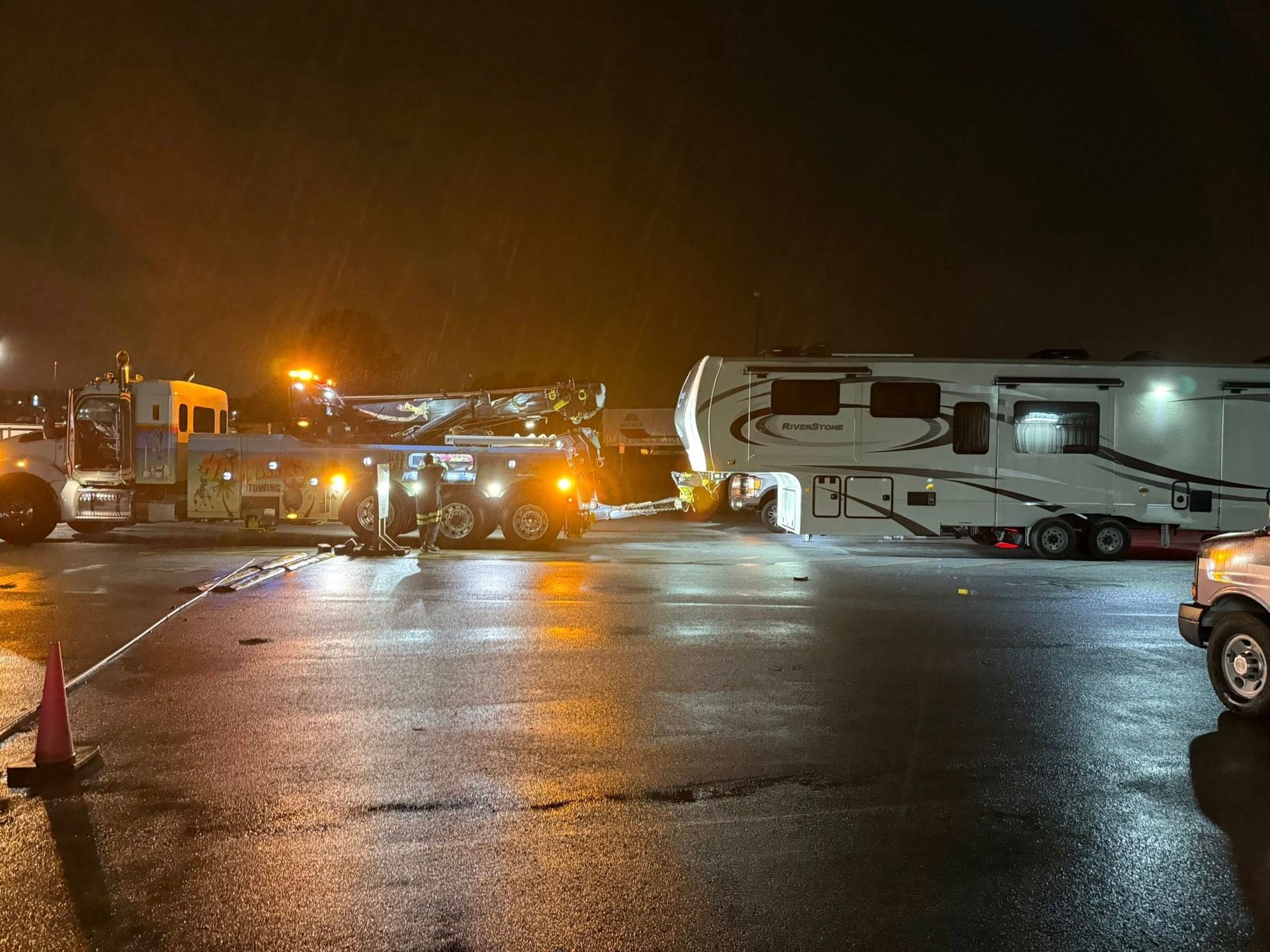 Tow truck and RV in a wet parking lot at night, with orange lights reflecting on the pavement