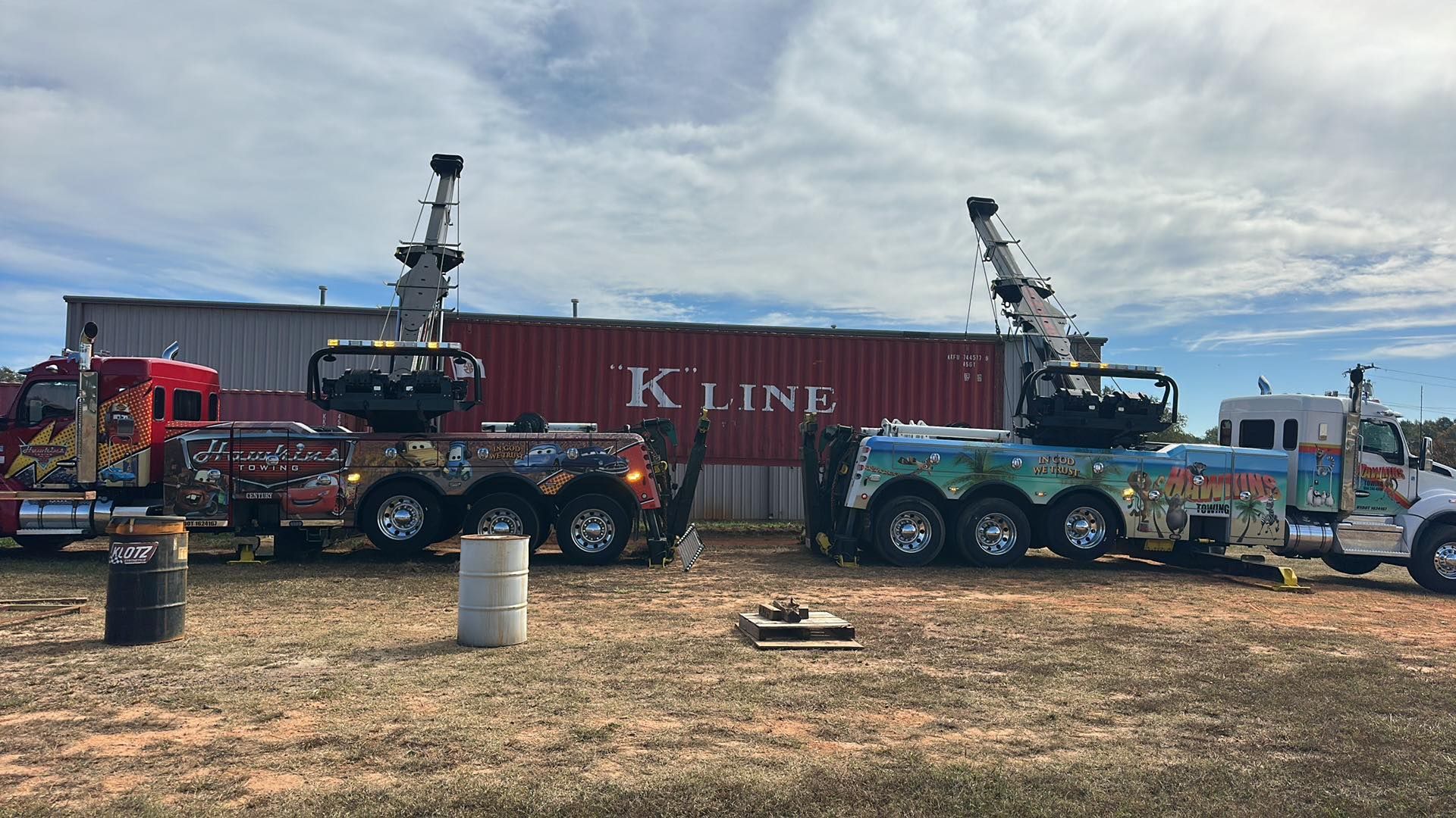 Two large tow trucks with mounted cranes parked in front of a K Line shipping container.