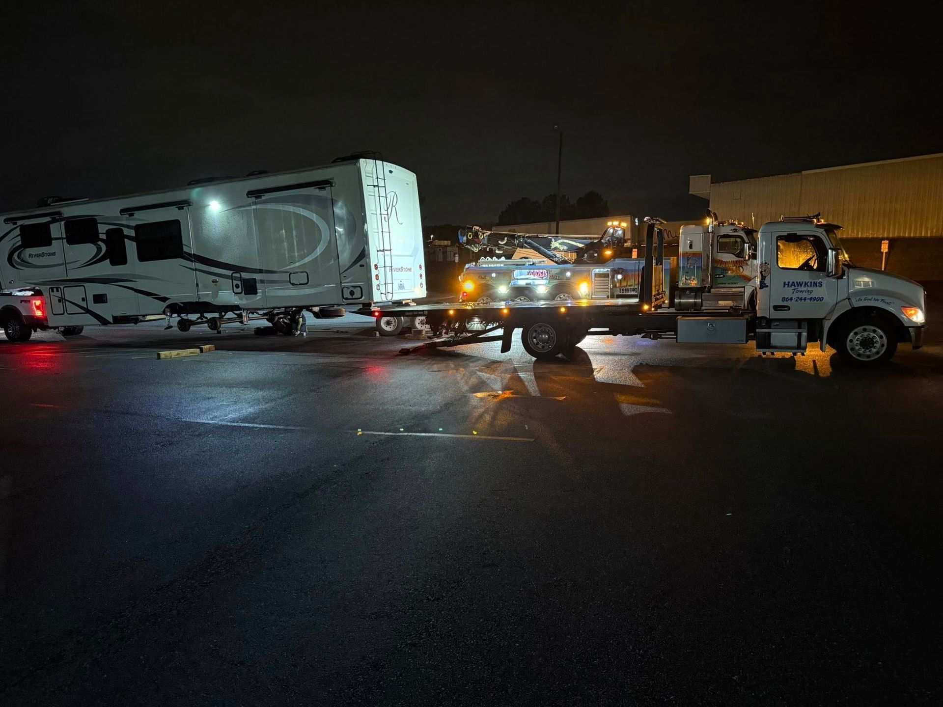 Tow truck lifting a trailer on a wet road at night, with headlights and city lights in the background