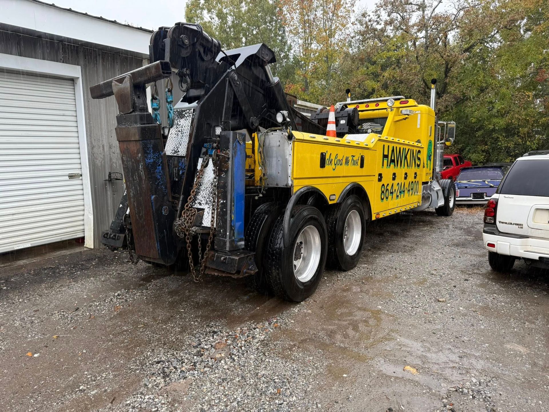 Yellow tow truck with a crane arm parked on a wet driveway beside a garage and white SUV