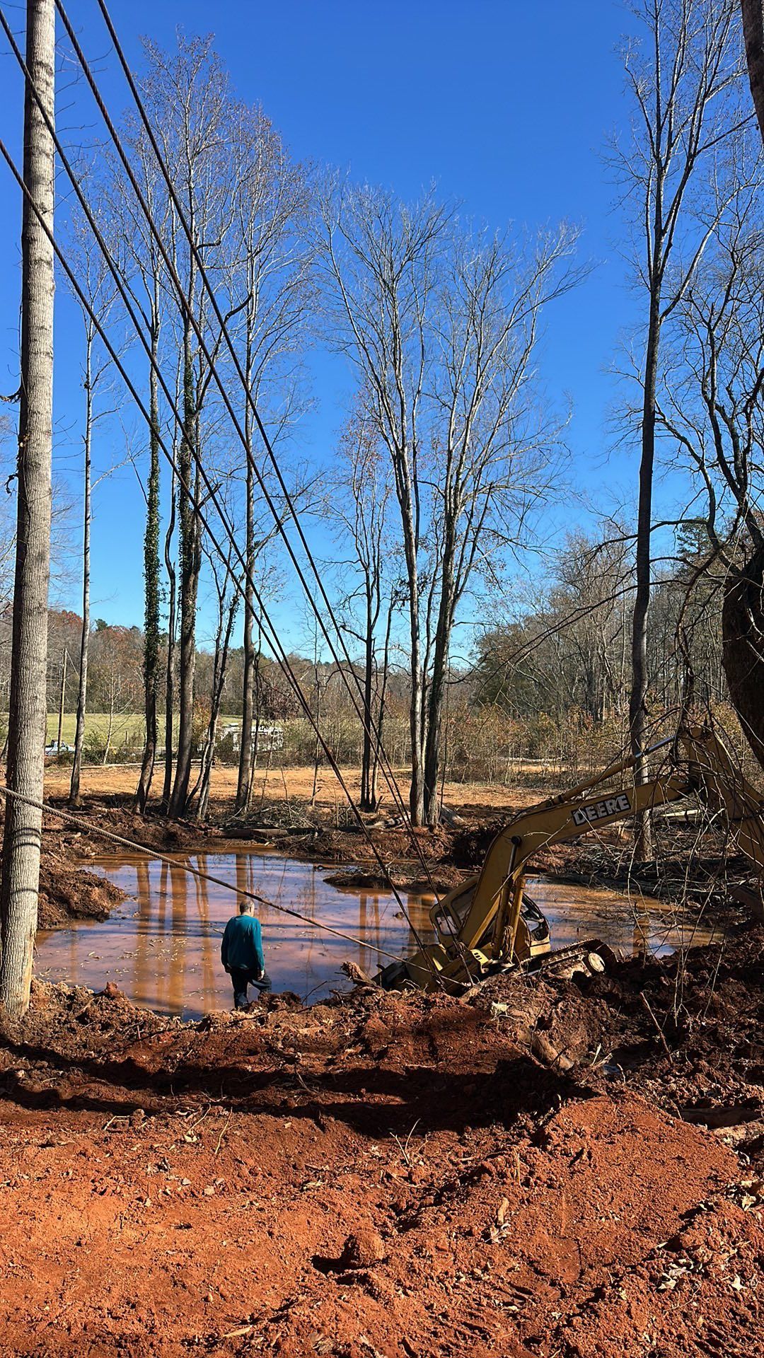 Small excavator digging muddy ground beside a pond in a leafless wooded area with one person standing nearby