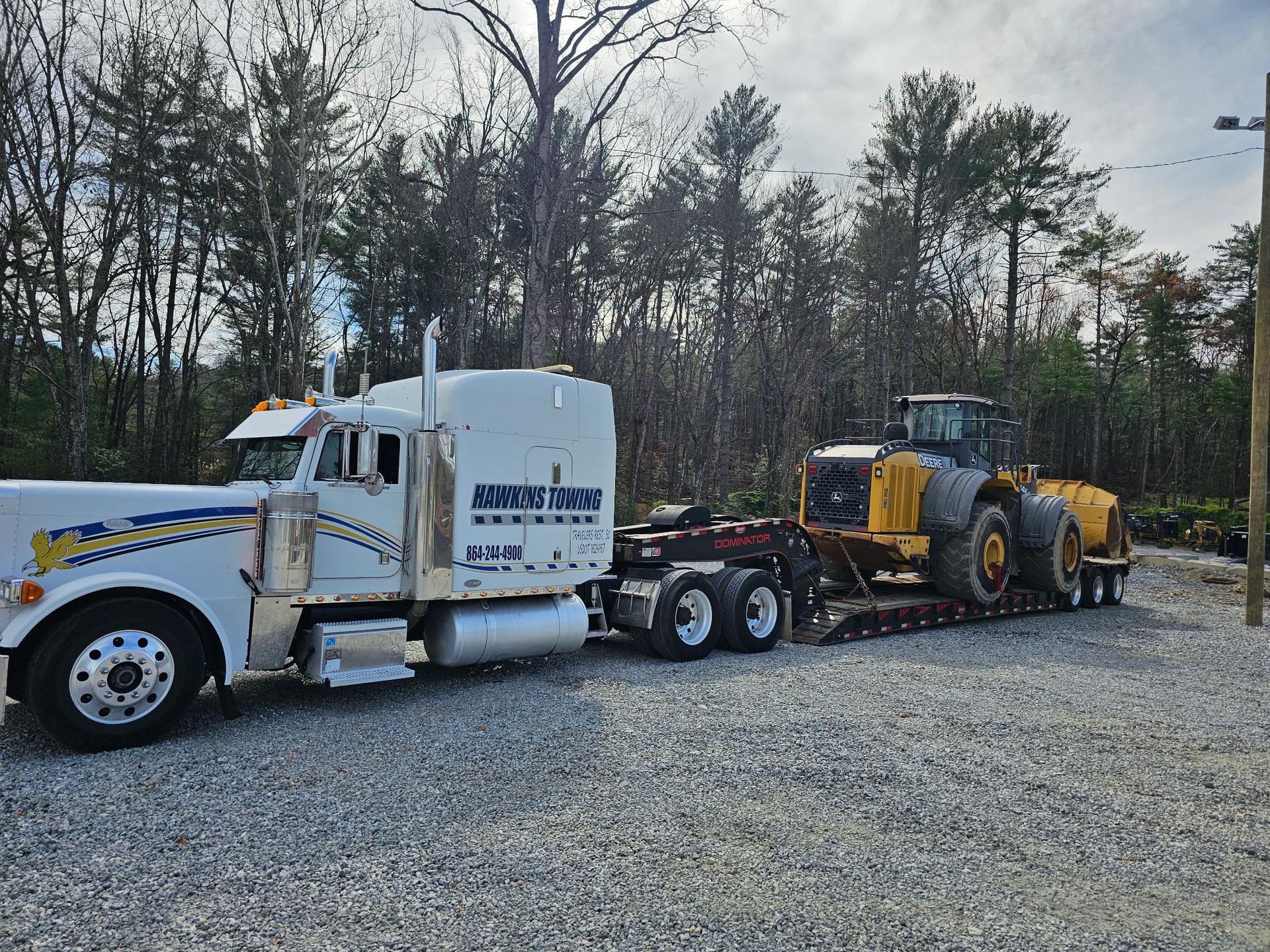 White semi-truck hauling a yellow tractor on a trailer in a gravel lot with trees behind