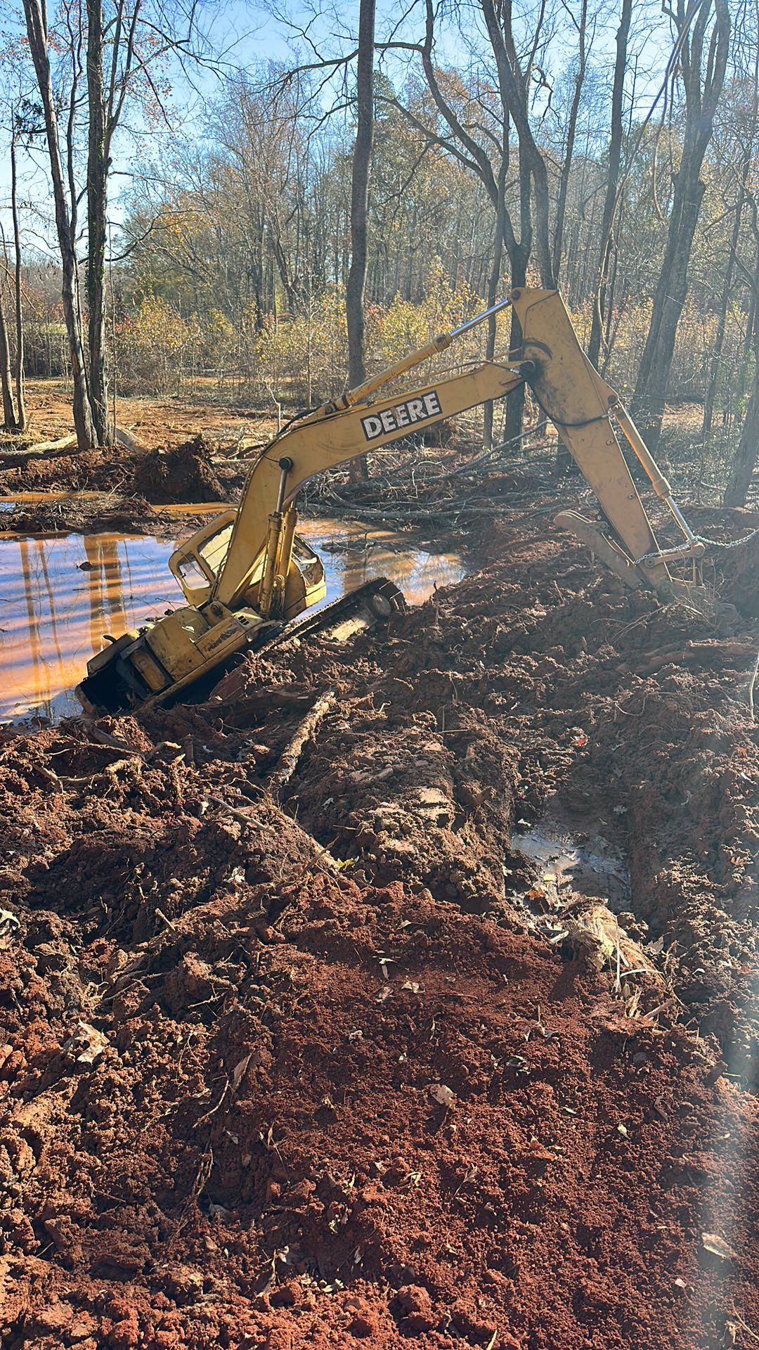 Yellow excavator digging a muddy pond edge in a wooded area with bare trees