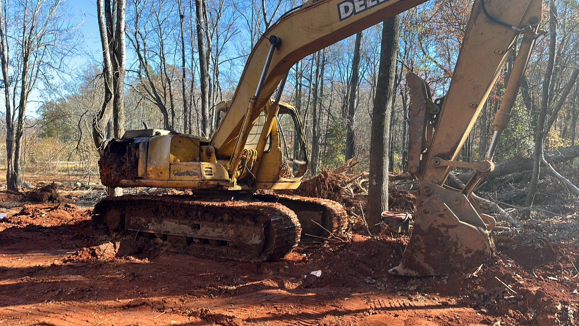 Yellow excavator working on a red dirt road in a leafless forest