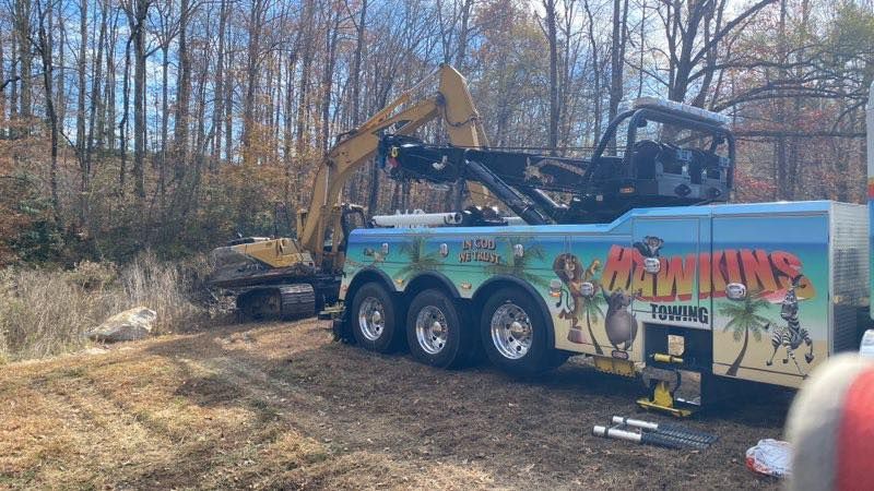 Tow truck hauling an excavator, with a colorful “Wreckers” trailer in a wooded area