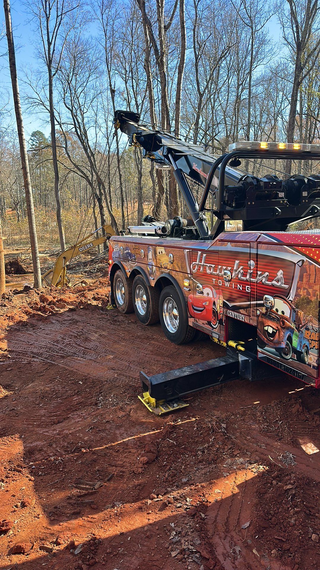 Red tow truck with boom arm parked on muddy dirt road in leafless wooded area